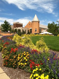 colorful flowers in front of a large building with grass and trees around the perimeter, on a sunny day