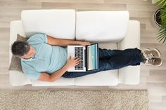 a man sitting on top of a white couch using a laptop computer next to a potted plant