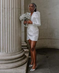 a woman standing next to a pillar with flowers in her hand and wearing a white dress