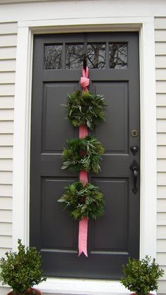 a wreath on the front door of a house with pink ribbon tied around it and two potted plants