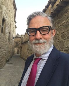 an older man wearing glasses and a suit standing in front of a stone building with cobblestone streets