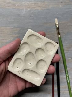 a hand holding a small white tray with six holes in it next to a paintbrush