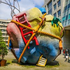 a giant rock with a ladder attached to it in front of a building and people walking by