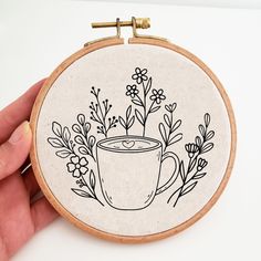 a hand holding a embroidery hoop with a cup of coffee and flowers in the middle