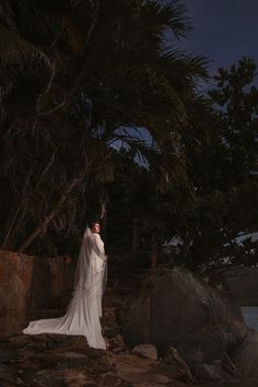 a woman in a wedding dress standing on rocks near the ocean at night with her arms around her head