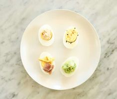 four deviled eggs on a white plate sitting on a marble counter top with different toppings