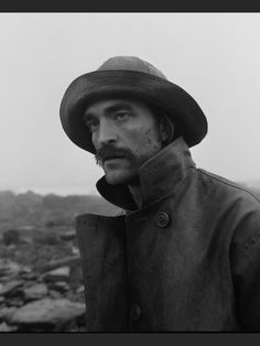 a man wearing a hat and trench coat standing in front of rocks on a cloudy day