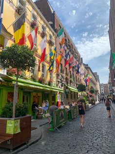 people are walking down the cobblestone street in front of buildings with flags on them