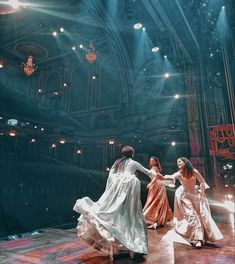 three women in long dresses are dancing on the stage with lights shining from behind them