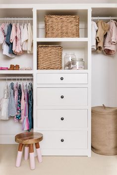 a white closet with clothes and baskets on the shelves, next to a wooden stool