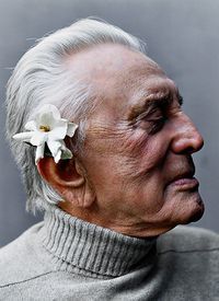 an old man with white hair and a flower in his ear is looking off to the side