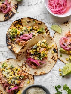 three tacos with corn, red onions and cilantro on the side next to some lime wedges