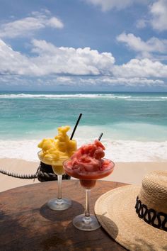 two cocktails are sitting on the beach with straw hats and blue water in the background