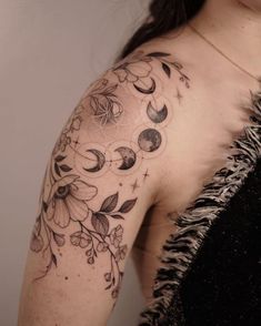 a woman's shoulder with flowers and moon tattoos on her left arm, behind her is a feathery black dress