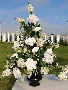 a black vase filled with white flowers sitting on top of a grass covered field next to a white picket fence