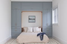 a bed sitting under a window in a bedroom next to blue cabinets and drawers with white pillows