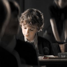 a young boy sitting at a desk in front of another person with his eyes closed