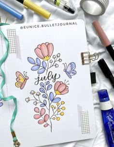 an open notebook with the words july surrounded by markers, pens and other office supplies