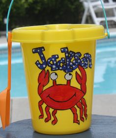 a yellow bucket with a crab on it next to a swimming pool and an umbrella