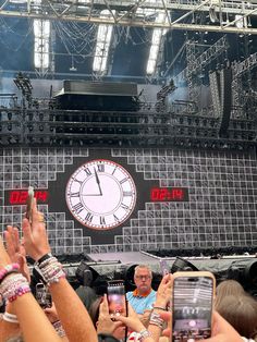 people taking pictures with their cell phones at an outdoor music festival in front of a giant clock