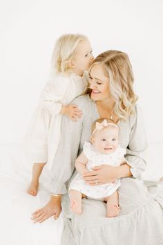 a woman holding a baby while sitting on top of a white bed next to another woman