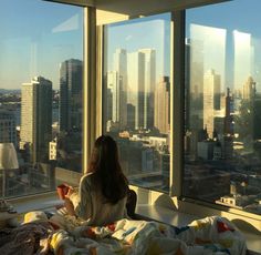 a woman sitting on top of a bed in front of a window looking out at the city