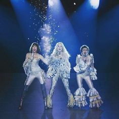 three women dressed in silver and white performing on stage with lights shining down behind them