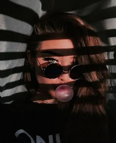 a woman with sunglasses blowing bubble in front of her face and shadows on the wall behind her