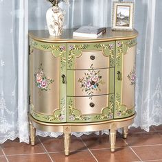 an ornate gold and green cabinet with flowers on the top, in front of a curtained window