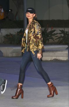 a woman in camouflage jacket and hat walking down street with her foot on the ground