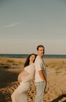 a pregnant couple standing on the beach in front of an ocean and sand dune area