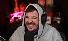 a man wearing headphones and a hoodie smiles while sitting in front of a microphone