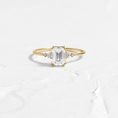 a gold ring with an emerald cut diamond and three small diamonds on the band, sitting on a white surface