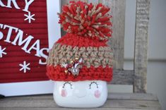 a red and beige knitted hat sitting on top of a white vase next to a christmas sign