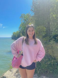 a woman in pink sweater and black shorts standing by the water with her hands on her hips