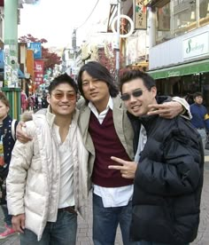 three young men standing next to each other on a city street with stores in the background