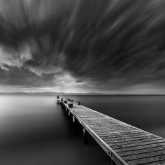 black and white photograph of a long pier in the ocean with storm clouds above it