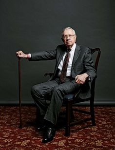 an older man in a suit and tie sitting on a chair with his arm extended