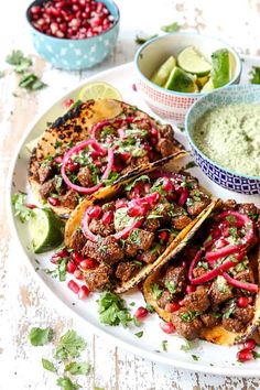 three tacos on a plate with guacamole, lime wedges and pomegranate