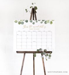 an easel with a calendar on it and greenery in the top right corner