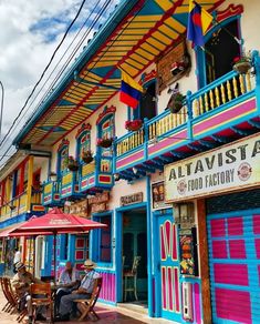 people sitting at tables in front of brightly colored buildings with awnings on the balconies