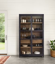 an open bookcase with dishes on it in front of a white wall and potted plant