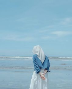 a woman in a white dress standing on the beach looking out at the ocean and holding her hands together