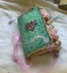 an old book is decorated with pink and green flowers, ribbons, and beads on a white tablecloth