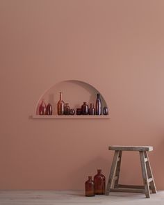 a shelf with vases and bottles on it next to a stool in front of a pink wall