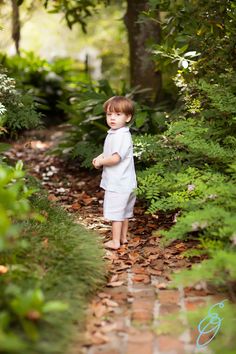 a little boy that is standing in the dirt with leaves on the ground and trees behind him