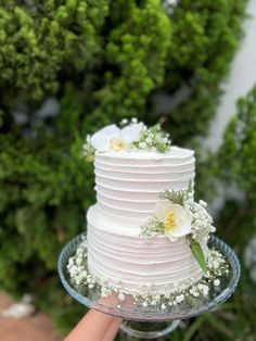 a white wedding cake with flowers and greenery on top is being held by a person