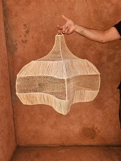 a person holding a woven object in front of a brown wall with a hand pointing at it