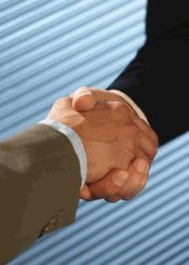 two men shaking hands in front of a blue building with corrugated roofing behind them