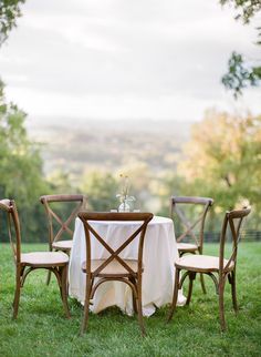 the table is set with four chairs and a white table cloth on it, in front of some trees